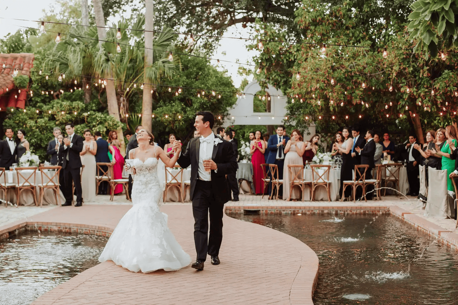 A bride and groom walk hand in hand on a brick path over a pond, surrounded by wedding guests clapping and watching under string lights in an outdoor garden setting at their wedding reception.