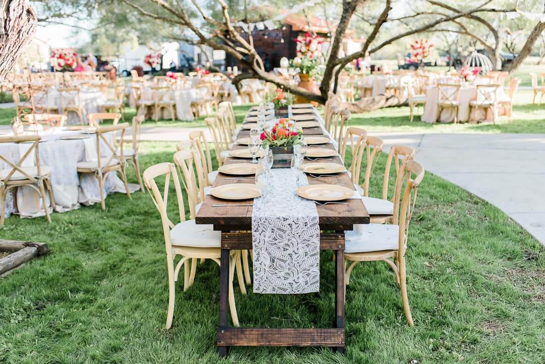 A long wooden table with a lace runner and simple floral centerpiece is set with plates and surrounded by wooden chairs at an outdoor wedding reception on a grassy lawn.