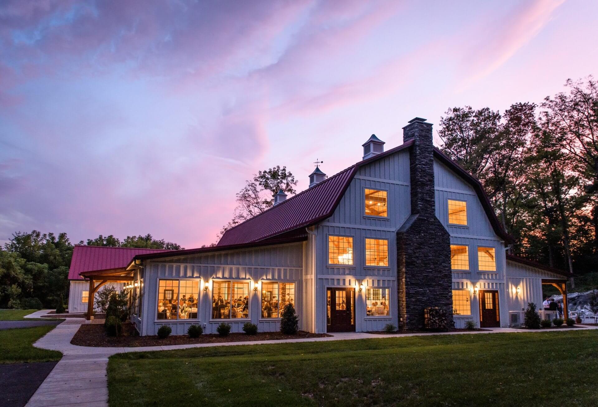 A large, modern barn-style building with illuminated windows and a stone chimney, perfect for a wedding reception, set against a sunset sky with trees in the background.