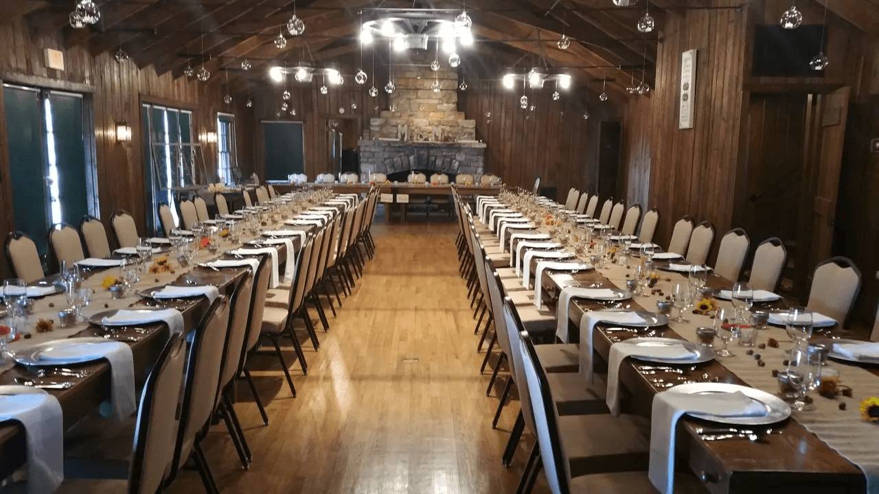Long dining tables set with plates, glasses, and napkins in a rustic wooden hall create a charming wedding reception atmosphere, complete with a stone fireplace and hanging lights.
