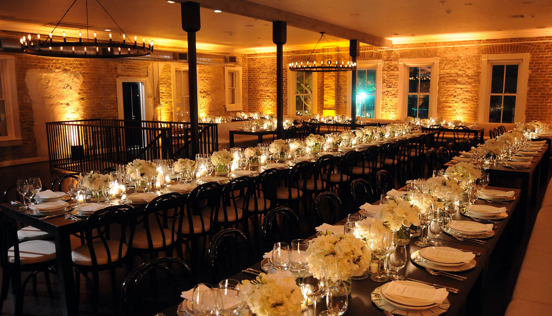 A banquet hall with long tables set for a wedding reception, featuring white floral centerpieces, glassware, plates, and candles, all illuminated by warm lighting.