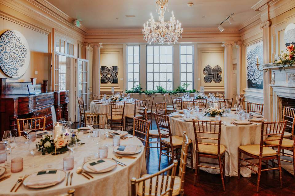 Elegant banquet room set for a wedding reception, featuring round tables, gold chairs, a sparkling chandelier, and large windows that bathe the space in natural light.
