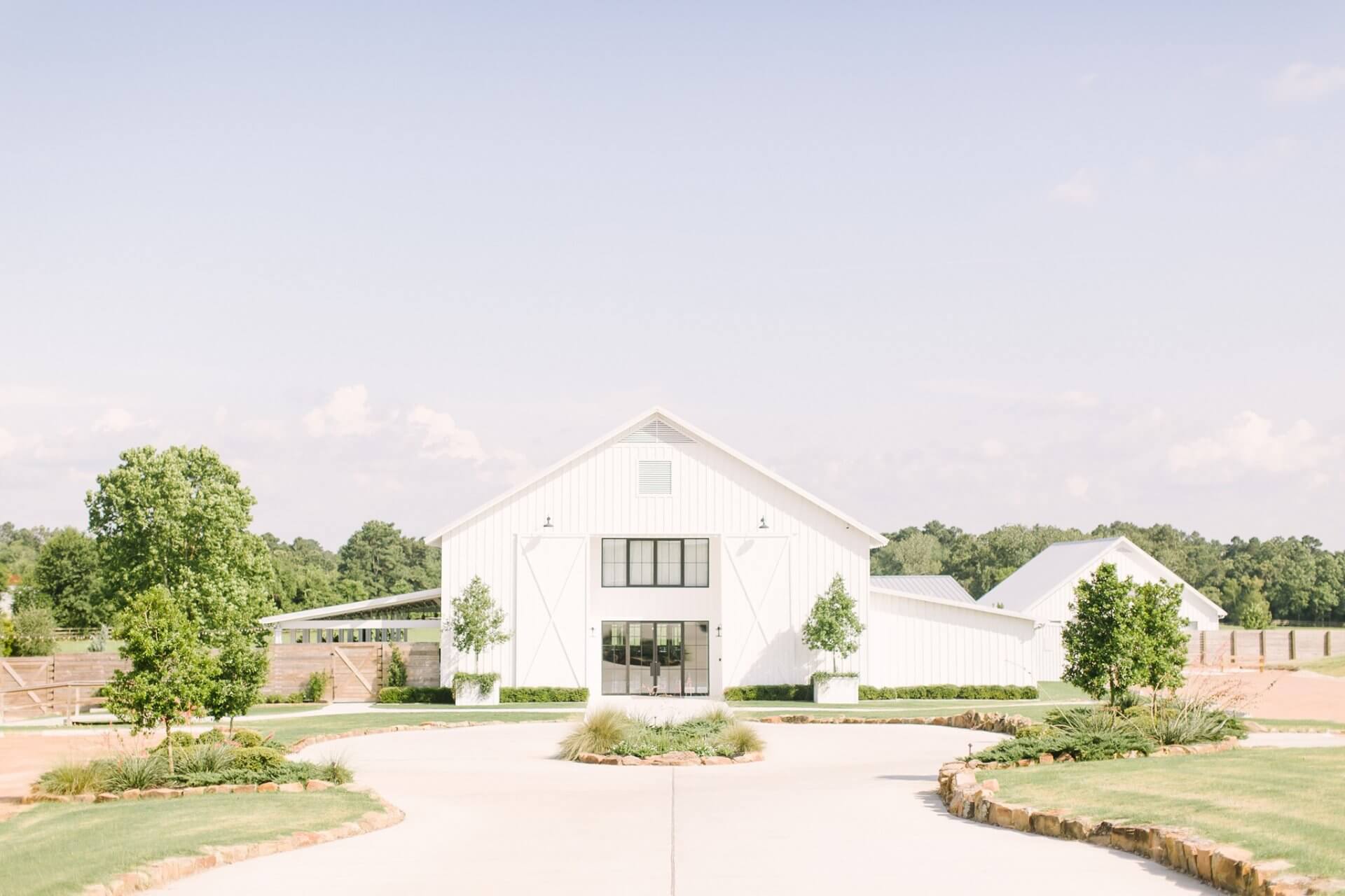 A large white barn with glass doors stands at the end of a curved driveway, perfect for a wedding reception, surrounded by green lawns, trees, and a clear sky.