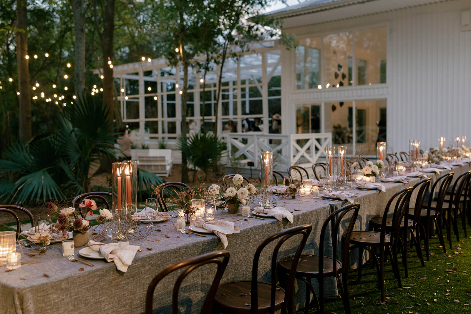 Long outdoor dining table set for a wedding reception with candles, flowers, and tableware, surrounded by wooden chairs near a white building with string lights in the background.