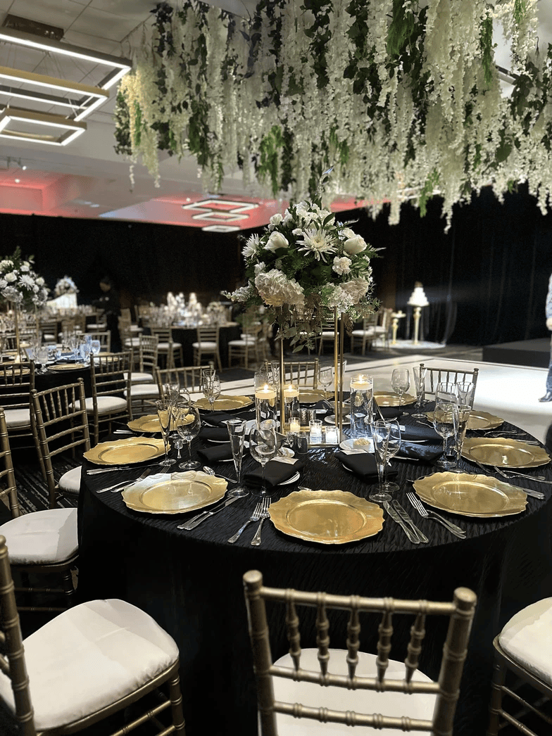 Round banquet table set with gold plates, black tablecloth, floral centerpiece, and candles for a stunning wedding reception; gold chairs surround the table beneath hanging greenery decor in an elegant event hall.