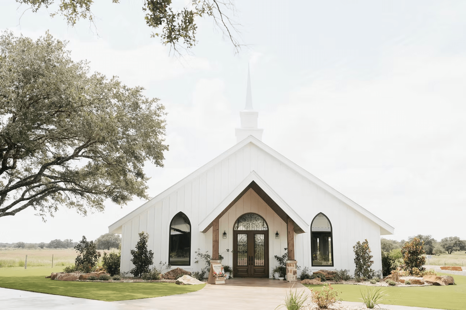 White church with a steeple, large windows, and double wooden doors, surrounded by landscaping and trees, set in a rural area—an ideal spot for a charming wedding reception under a cloudy sky.