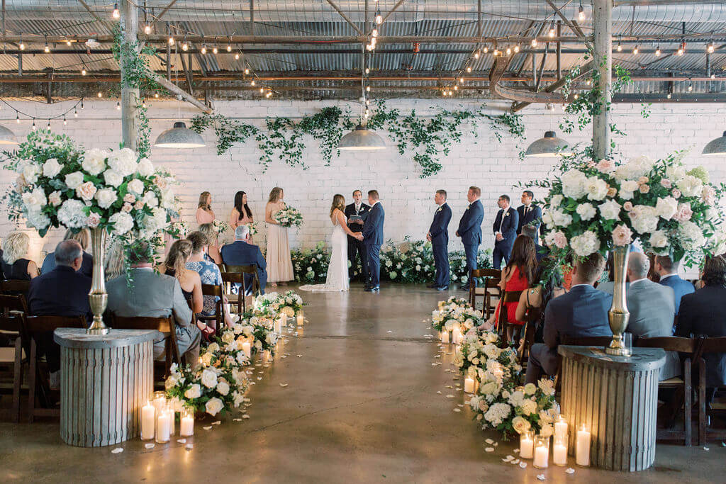 A couple stands at the altar in an industrial-style venue, surrounded by their wedding party, floral arrangements, and guests seated on either side of a candle-lined aisle—one of the most stunning wedding venues in Fort Worth.