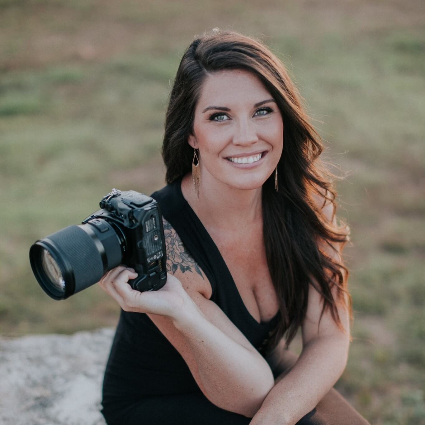 A woman with long brown hair and a black top sits outdoors holding a DSLR camera, smiling at the camera.