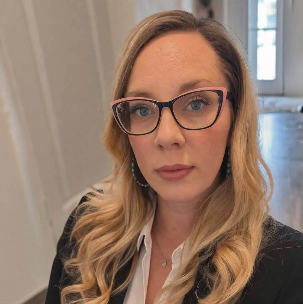 A woman with long blonde hair and glasses wearing a black blazer and white shirt stands indoors, looking at the camera.