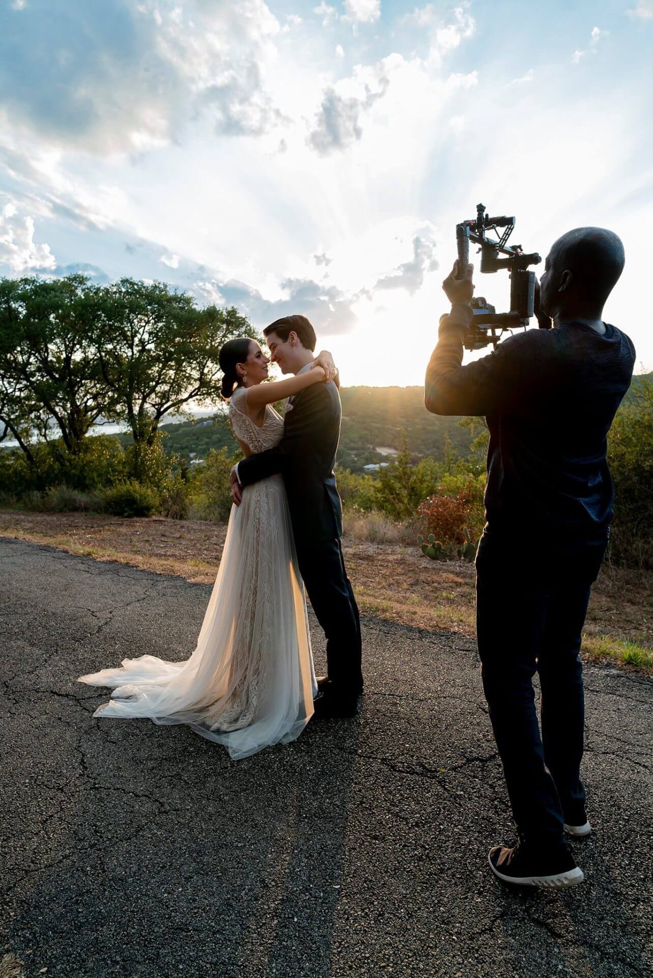 A bride and groom embrace outdoors at sunset while a videographer records them with a camera rig on a rural road.