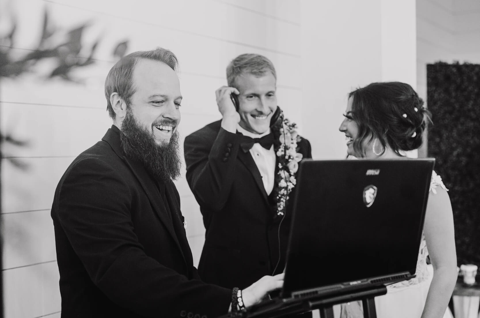 Three people dressed formally stand by a laptop; one man with a beard smiles at the screen while a couple in wedding attire looks on.