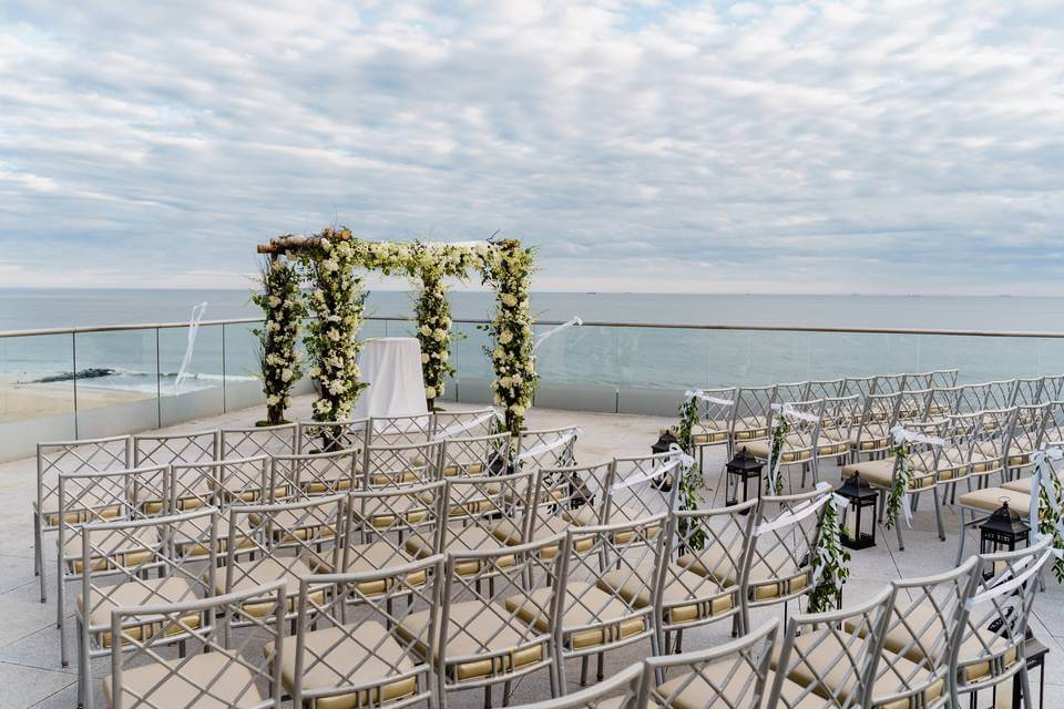 Rows of chairs face a floral wedding arch on a terrace overlooking the ocean, with cloudy skies and calm water—an ambiance reminiscent of top wedding venues in Austin.