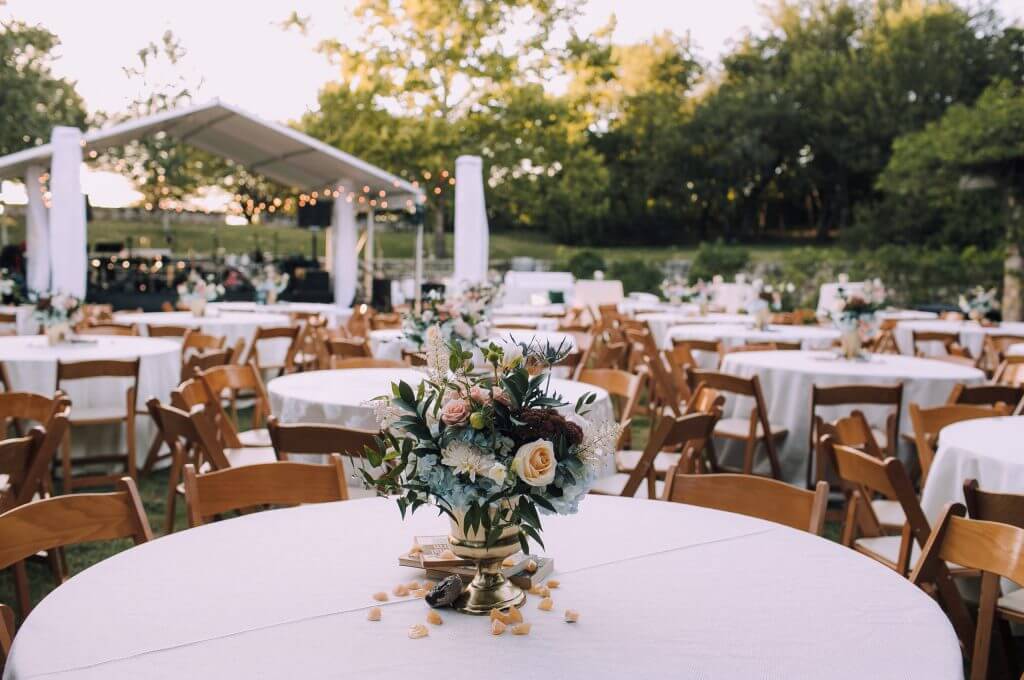 Outdoor event setup featuring round tables draped in white cloths, wooden chairs, and floral centerpieces; a tent with string lights glows in the background—an elegant scene perfect for San Antonio wedding venues.