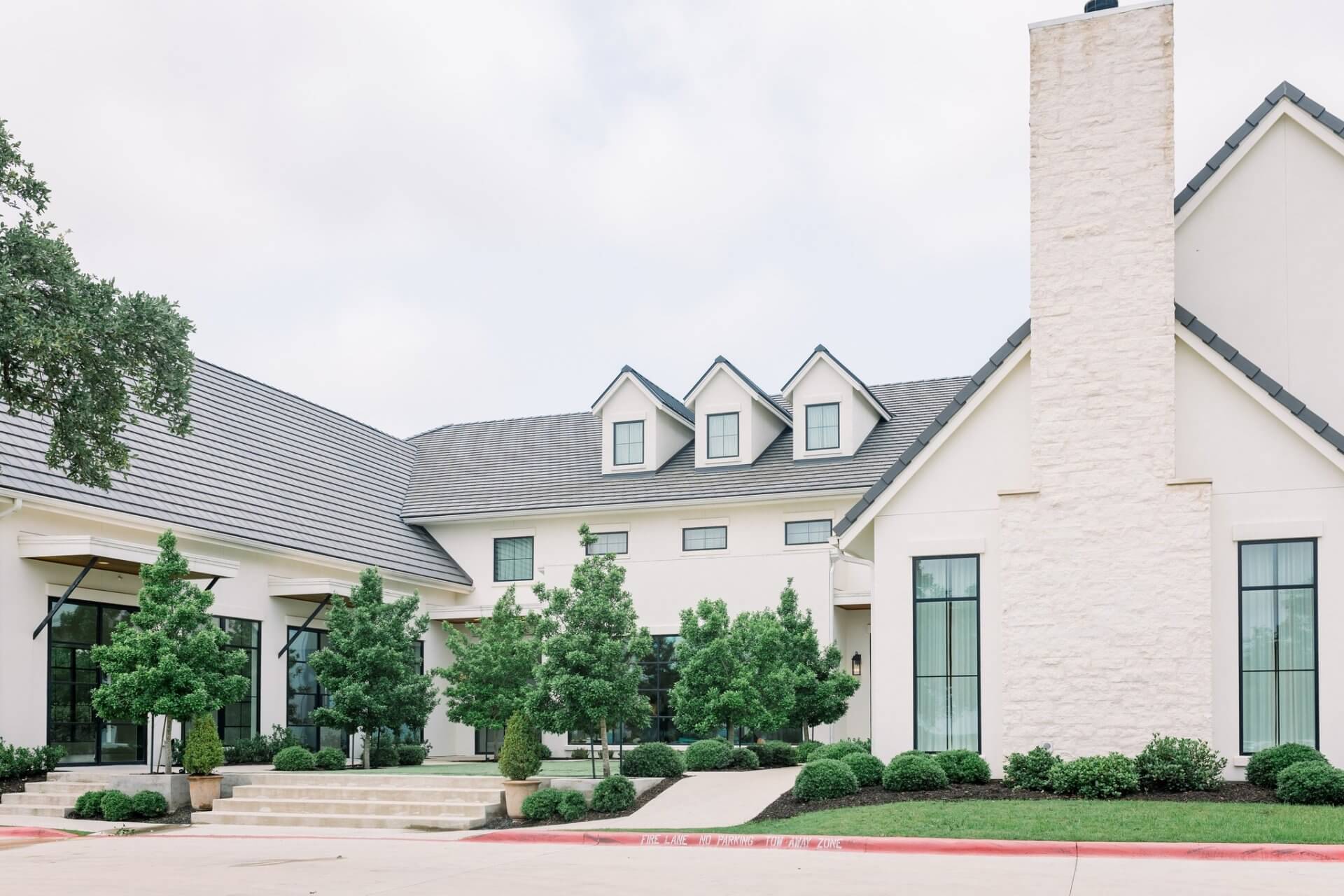 Modern white two-story house with large windows, dormer windows on the roof, a tall chimney, manicured shrubs, and a paved driveway—perfect for those seeking elegant wedding venues in Austin.