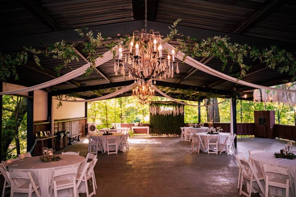 A decorated event space with round white-clothed tables, white chairs, draped fabric, chandeliers, and greenery, set under an open metal-roof pavilion with large windows—one of the most elegant wedding venues in Fort Worth.
