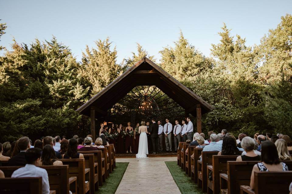 A wedding ceremony takes place outdoors under a wooden pavilion at one of the elegant wedding venues in Fort Worth, with the bride and groom standing at the altar and guests seated in rows.
