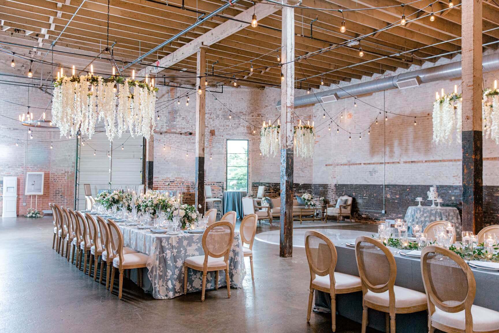 A decorated event space with long banquet tables, elegant chairs, floral centerpieces, hanging chandeliers, and exposed brick walls beneath high wooden ceilings—perfect for those seeking unique wedding venues in Fort Worth.