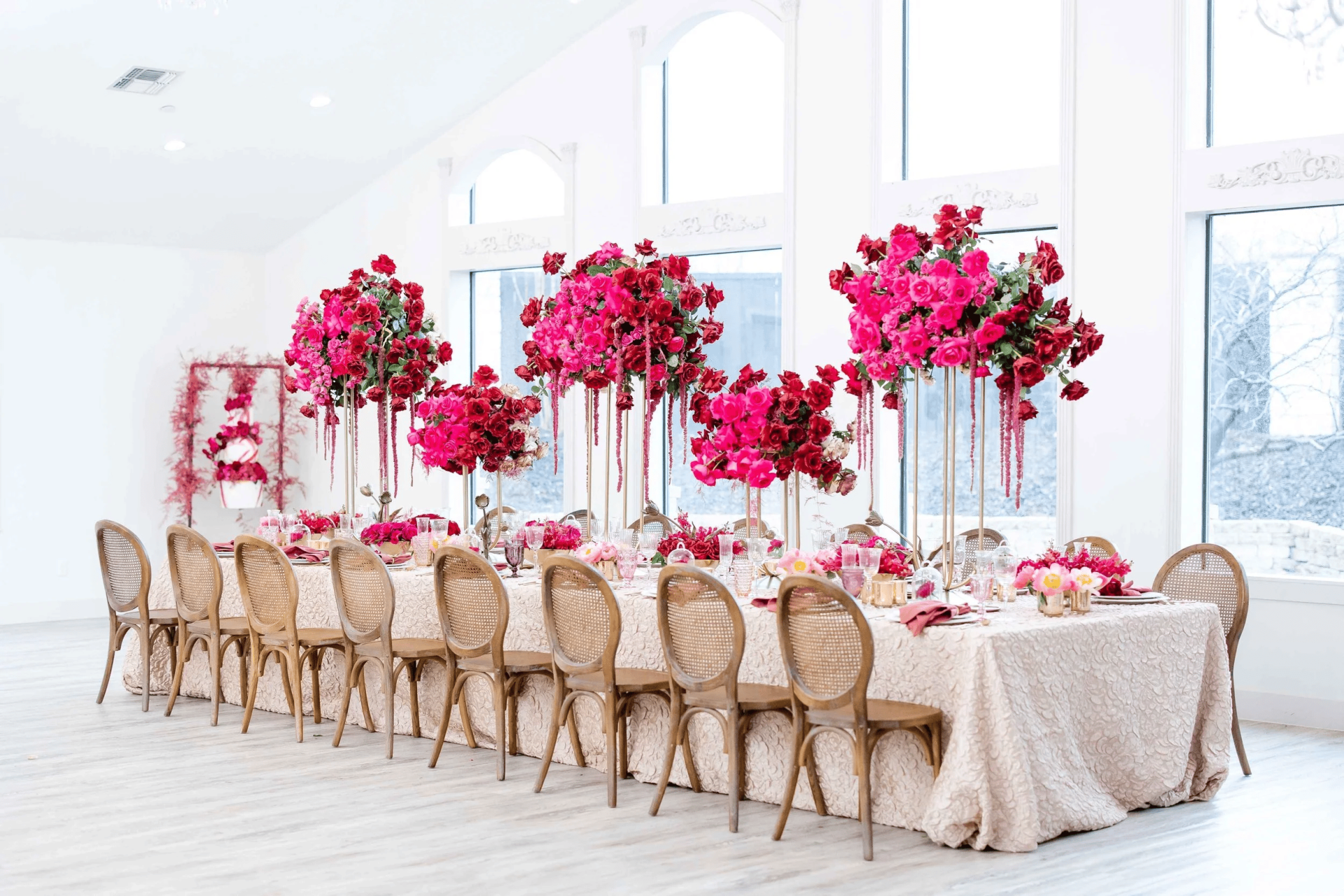 A long banquet table with beige fabric, wooden chairs, and tall floral centerpieces of bright pink and red flowers in a bright room with large windows—perfect inspiration for wedding venues in Fort Worth.