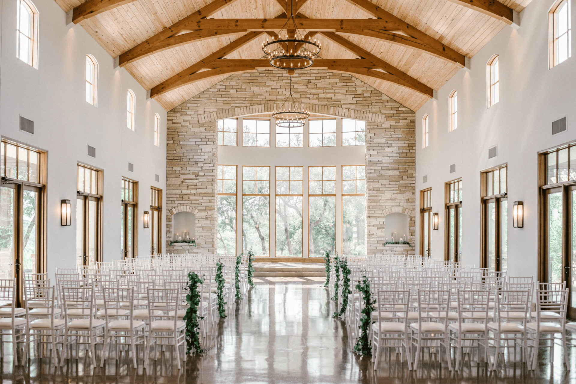 A spacious, light-filled wedding venue in Austin features rows of white chairs with green aisle decorations, wooden beams on the ceiling, and large windows overlooking trees.