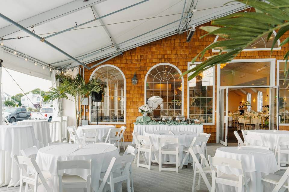 Outdoor event space with round tables and white chairs set up under a tent, in front of a building with arched windows and wood siding; one of the elegant wedding venues in Austin, decorated with white tablecloths and flowers.