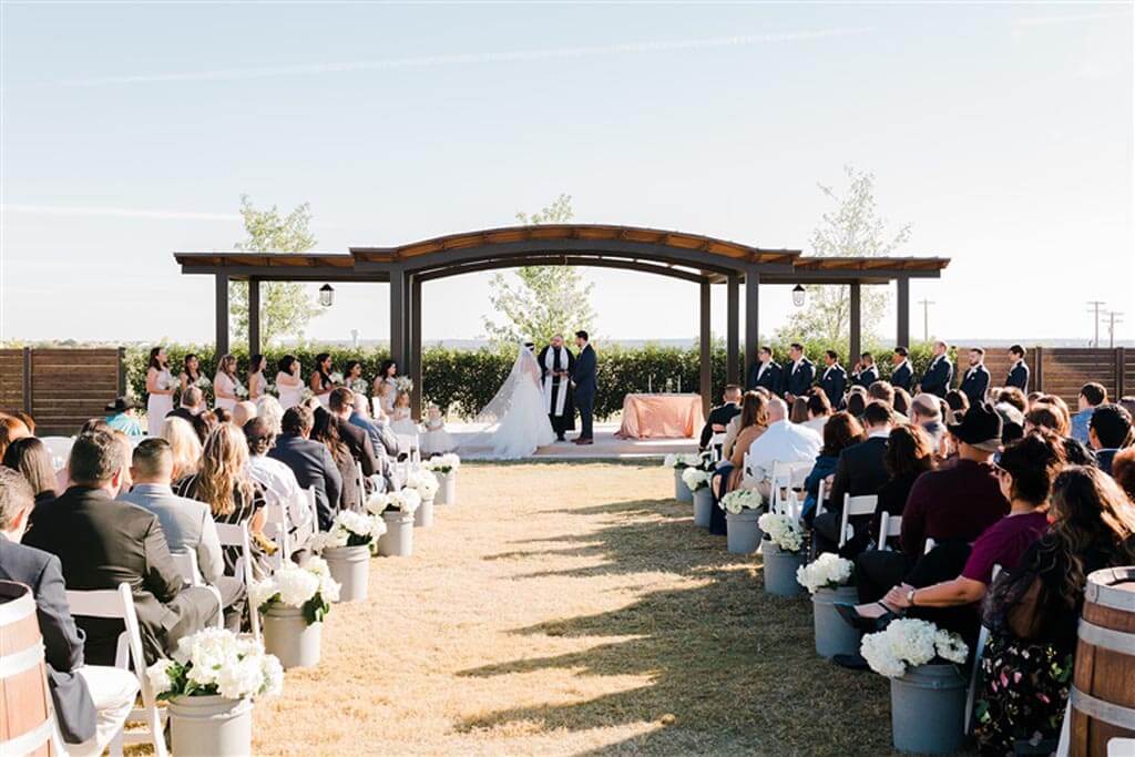 Outdoor wedding ceremony with bride and groom standing under an arch, surrounded by their wedding party and guests, with flower arrangements lining the aisle—perfect for those seeking beautiful wedding venues in Austin.