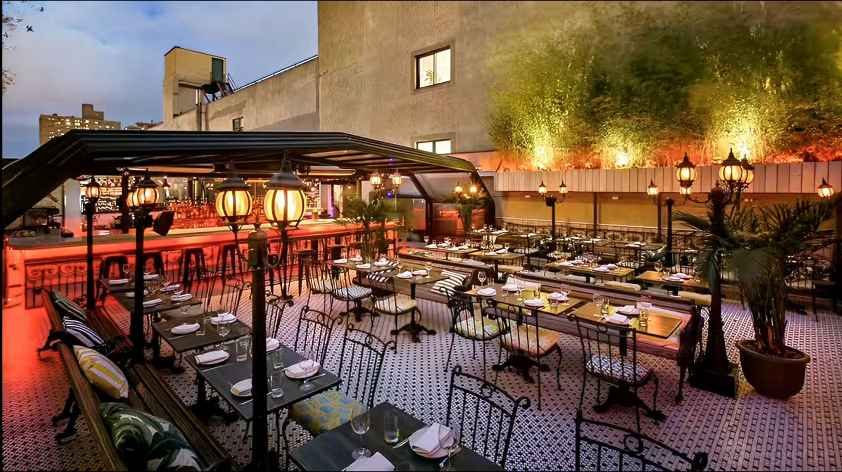Outdoor restaurant patio with black metal tables and chairs, covered bar area, decorative lamps, and plants along the lit wall—perfect for relaxed evenings or exploring unique wedding venues in Austin.