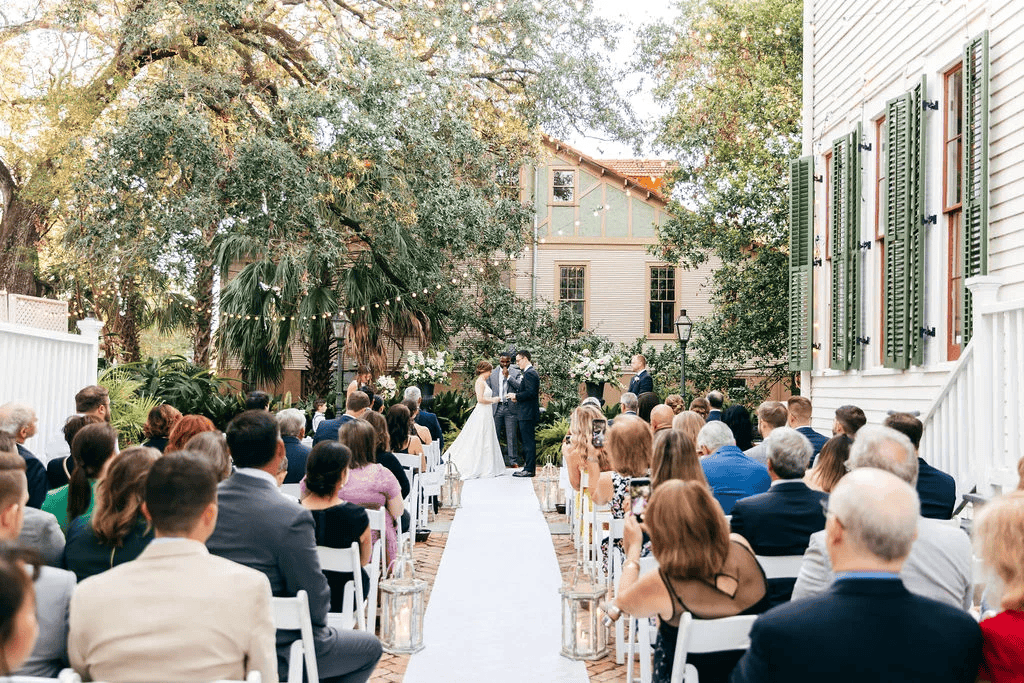 A wedding ceremony takes place outdoors at one of the charming New Orleans wedding venues, with guests seated on either side of a white aisle; the couple stands at the altar in front of an officiant under trees and string lights.