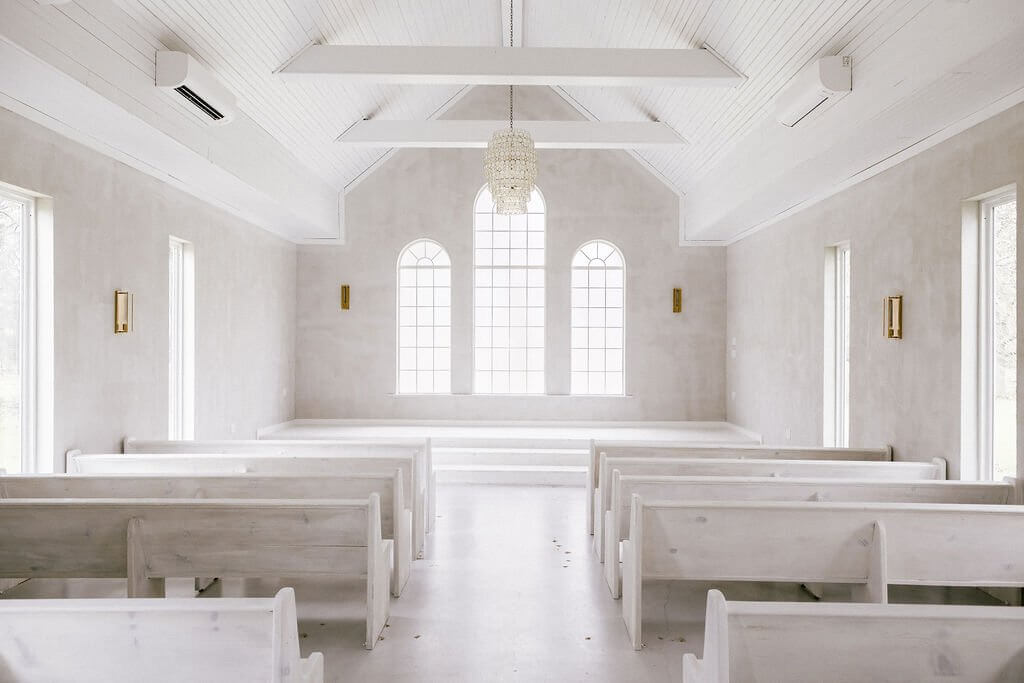 Bright, empty chapel interior with white wooden benches, tall arched windows, white walls, and a chandelier hanging from the ceiling—perfect for those seeking elegant wedding venues in Fort Worth.