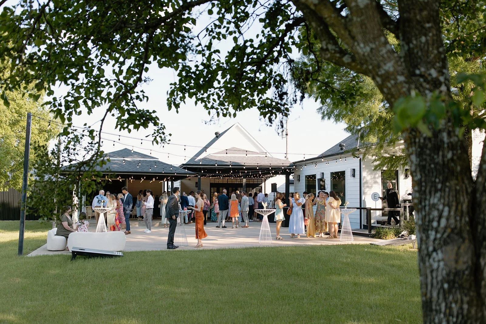 A group of people socialize outdoors at a daytime event near a white building, with string lights overhead and trees in the foreground—an inviting scene often found at wedding venues in Fort Worth.