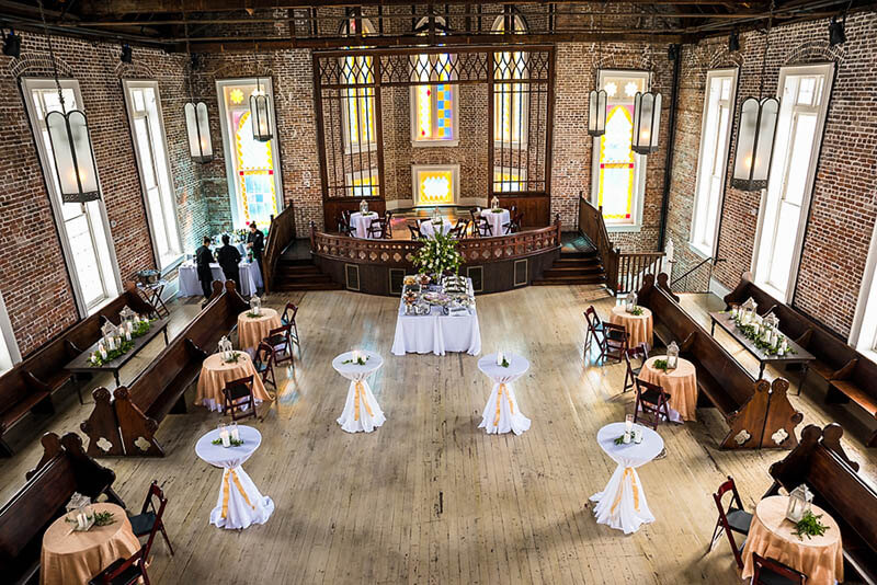 A historic brick-walled hall with stained glass windows, wooden pews, and round tables arranged for an event—perfect for those seeking unique New Orleans wedding venues, with a buffet table as the centerpiece.