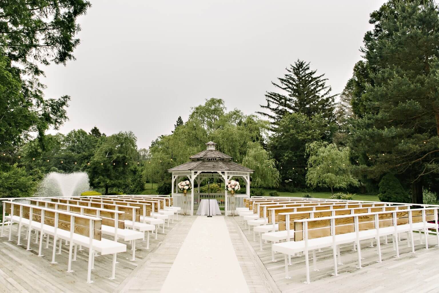 Outdoor wedding venue with white benches facing a flower-adorned gazebo, surrounded by trees and greenery, and a fountain on the left—an enchanting choice for those seeking premier wedding venues in Austin.