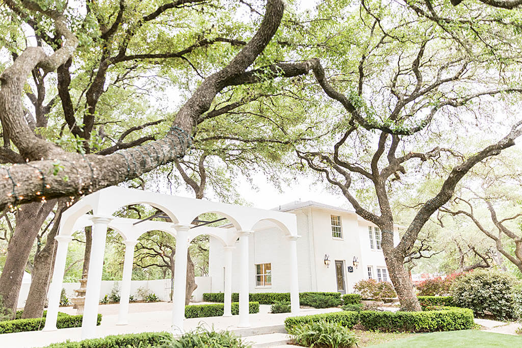 A white two-story house with an arched pergola is surrounded by large, sprawling trees and neatly trimmed bushes, creating an inviting setting perfect for those seeking charming San Antonio wedding venues.