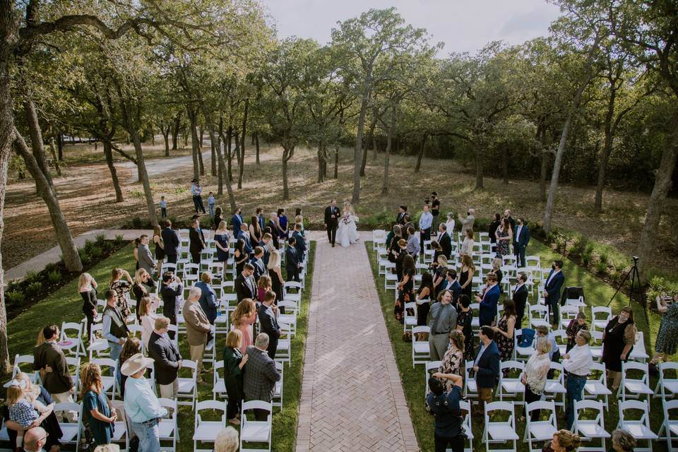 Outdoor wedding ceremony with rows of white chairs, guests standing on either side of a brick aisle as a couple stands at the altar surrounded by trees—perfect for those seeking picturesque wedding venues in Dallas.