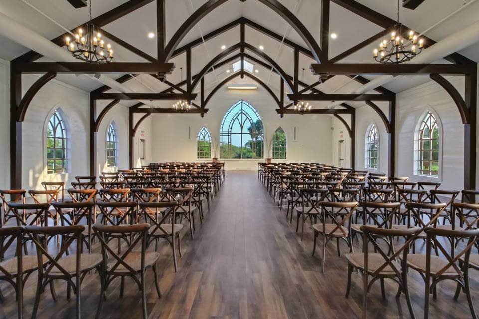 A bright, spacious hall with wooden floors, arched windows, and chandeliers—one of the most charming wedding venues in Austin—features rows of wooden chairs and exposed beams, all arranged for an event facing a small stage.