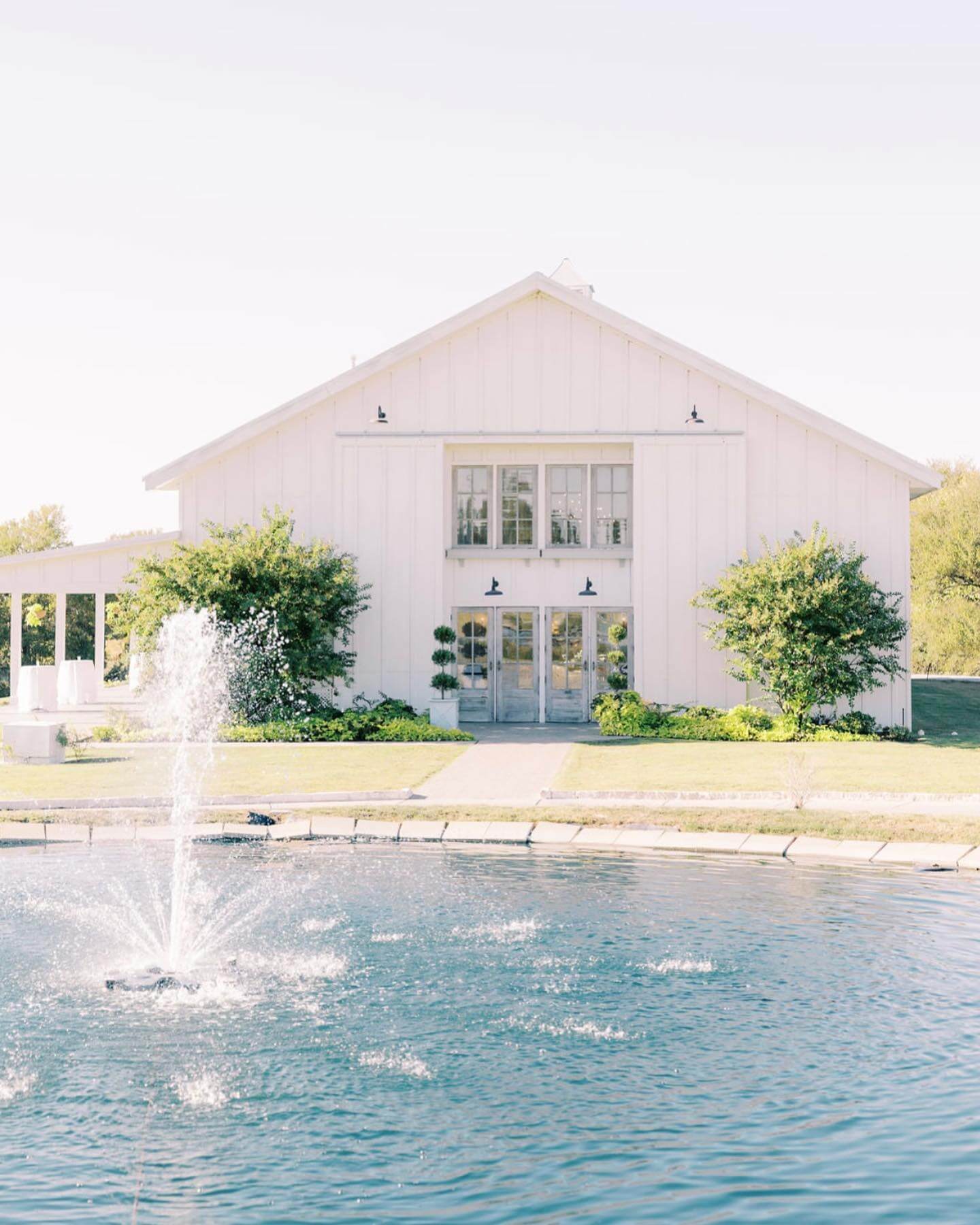 A white barn-style building with large windows stands behind a pond with a water fountain, surrounded by greenery under a clear sky—one of the charming wedding venues in Fort Worth.