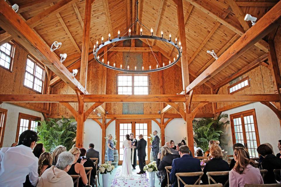 A wedding ceremony taking place in a wooden barn venue with high ceilings, large windows, and guests seated facing the couple and officiant—one of the most charming wedding venues in Austin.