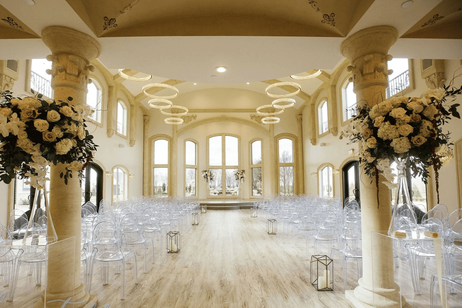 A bright, elegant indoor wedding venue in Fort Worth with tall arched windows, floral arrangements, transparent chairs, and decorative light rings hanging from the ceiling. Perfect for couples seeking stunning wedding venues in Fort Worth.