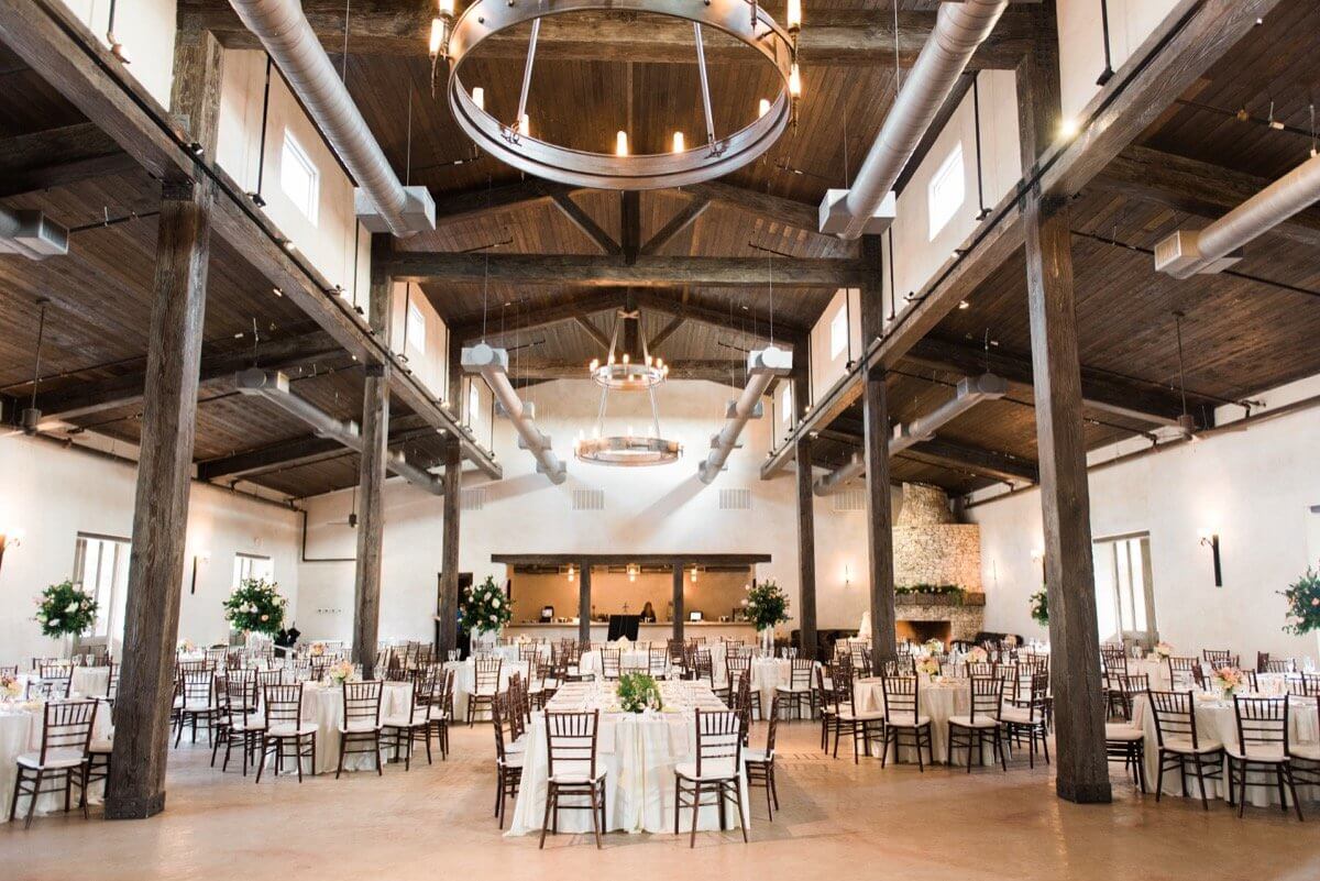Spacious banquet hall with wooden beams, chandeliers, round tables with white tablecloths, and arranged chairs—ideal for events and those searching for elegant San Antonio wedding venues.