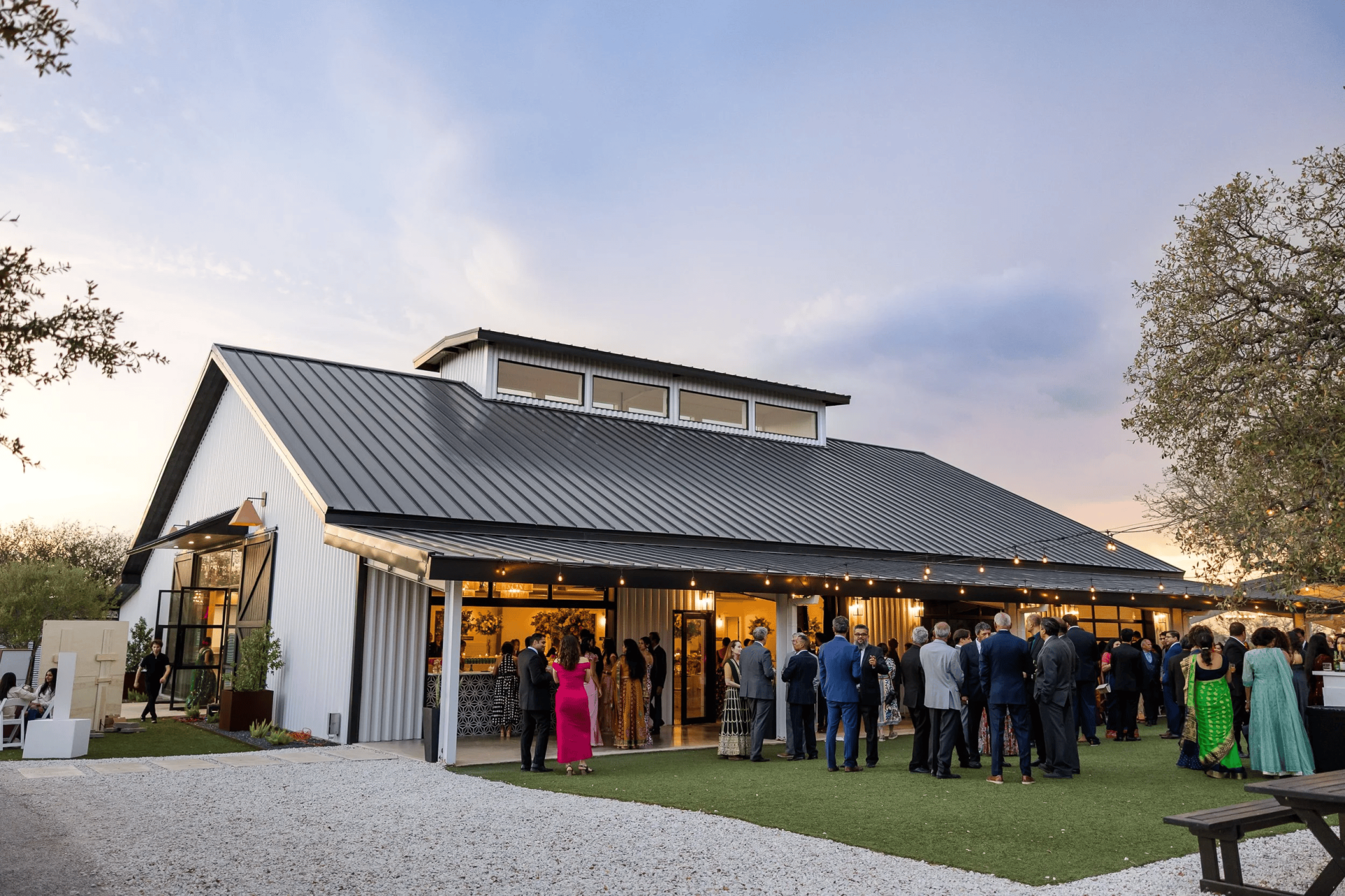 A group of people gather outside a modern barn-style venue at sunset, dressed in formal and semi-formal attire, with string lights overhead—a perfect scene for those seeking unique wedding venues in Austin.
