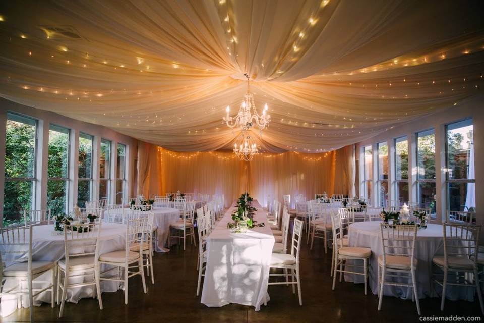 A banquet hall decorated with draped white fabric, chandeliers, string lights, round tables covered in white cloths, and white chairs arranged for an event—perfect for those seeking elegant wedding venues in Fort Worth.