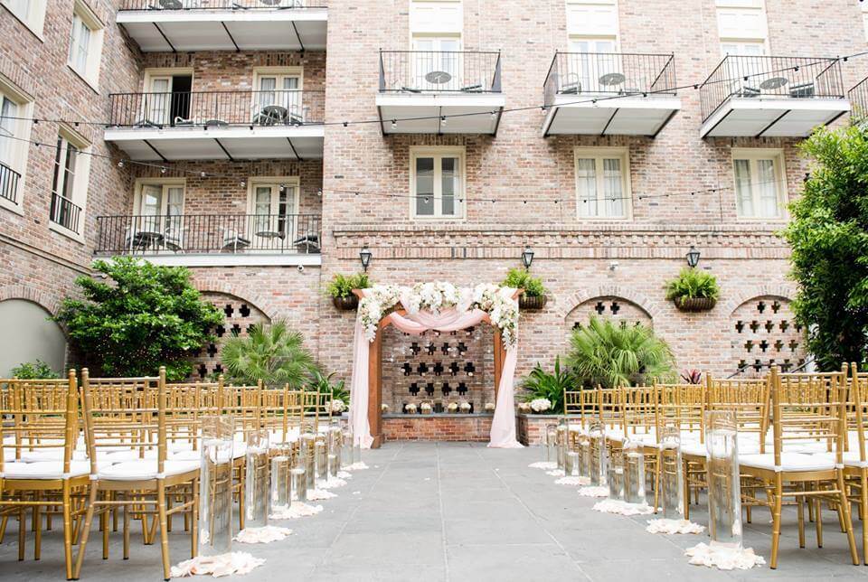 Outdoor wedding ceremony setup with gold chairs facing a floral and fabric-decorated arch, set in a courtyard with brick walls and balconies—perfect for those seeking enchanting New Orleans wedding venues.