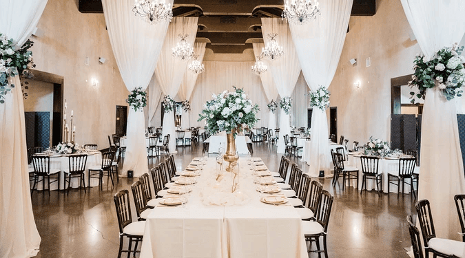 Elegant banquet hall with draped white curtains, floral centerpieces, round and rectangular tables set with plates, and chandeliers hanging from the ceiling—an ideal choice among wedding venues in Dallas.