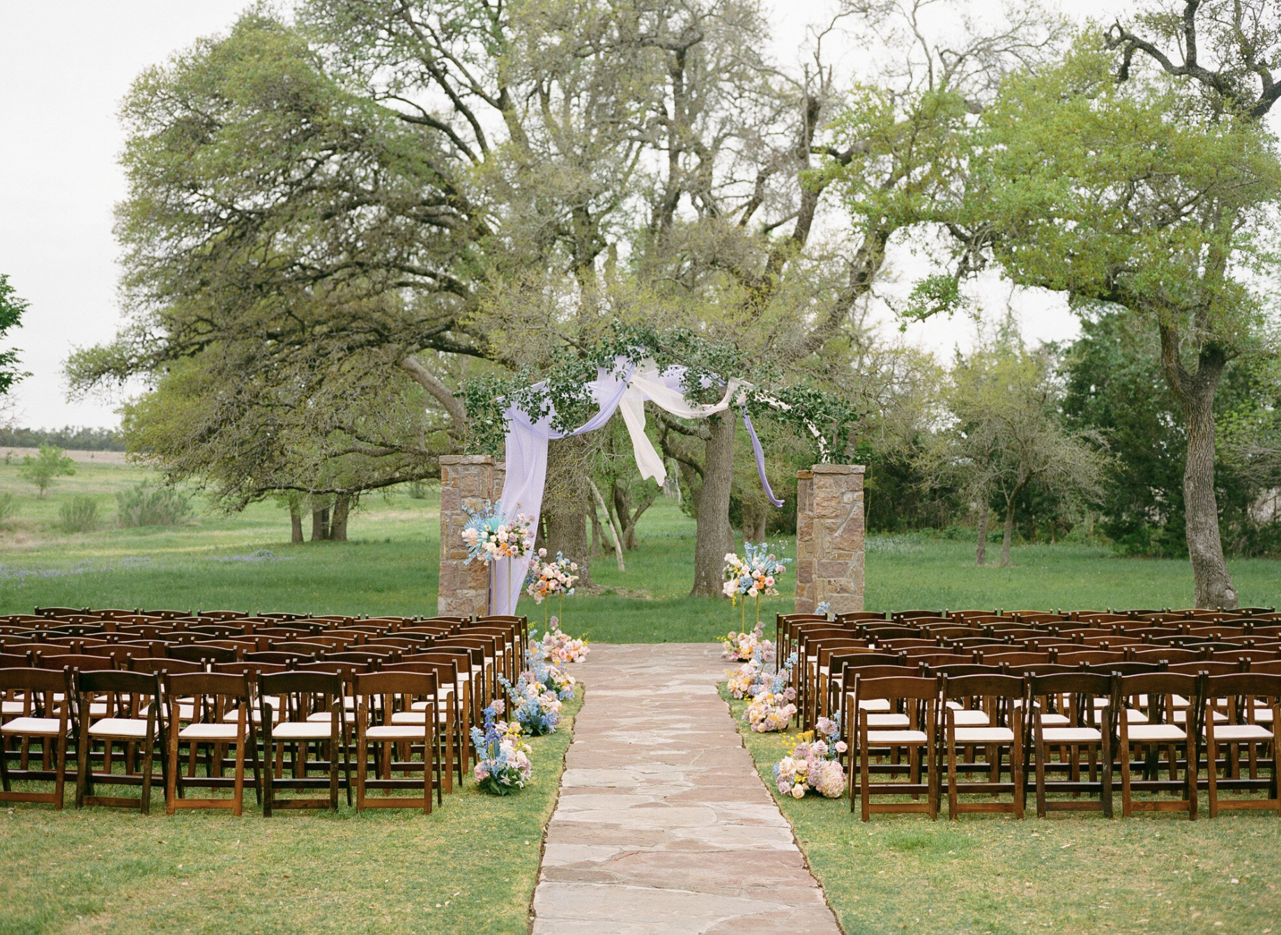 Outdoor wedding ceremony setup with rows of wooden chairs facing a stone arch adorned with flowers and flowing fabric, surrounded by green trees and grass—perfect inspiration for those exploring wedding venues in Dallas.