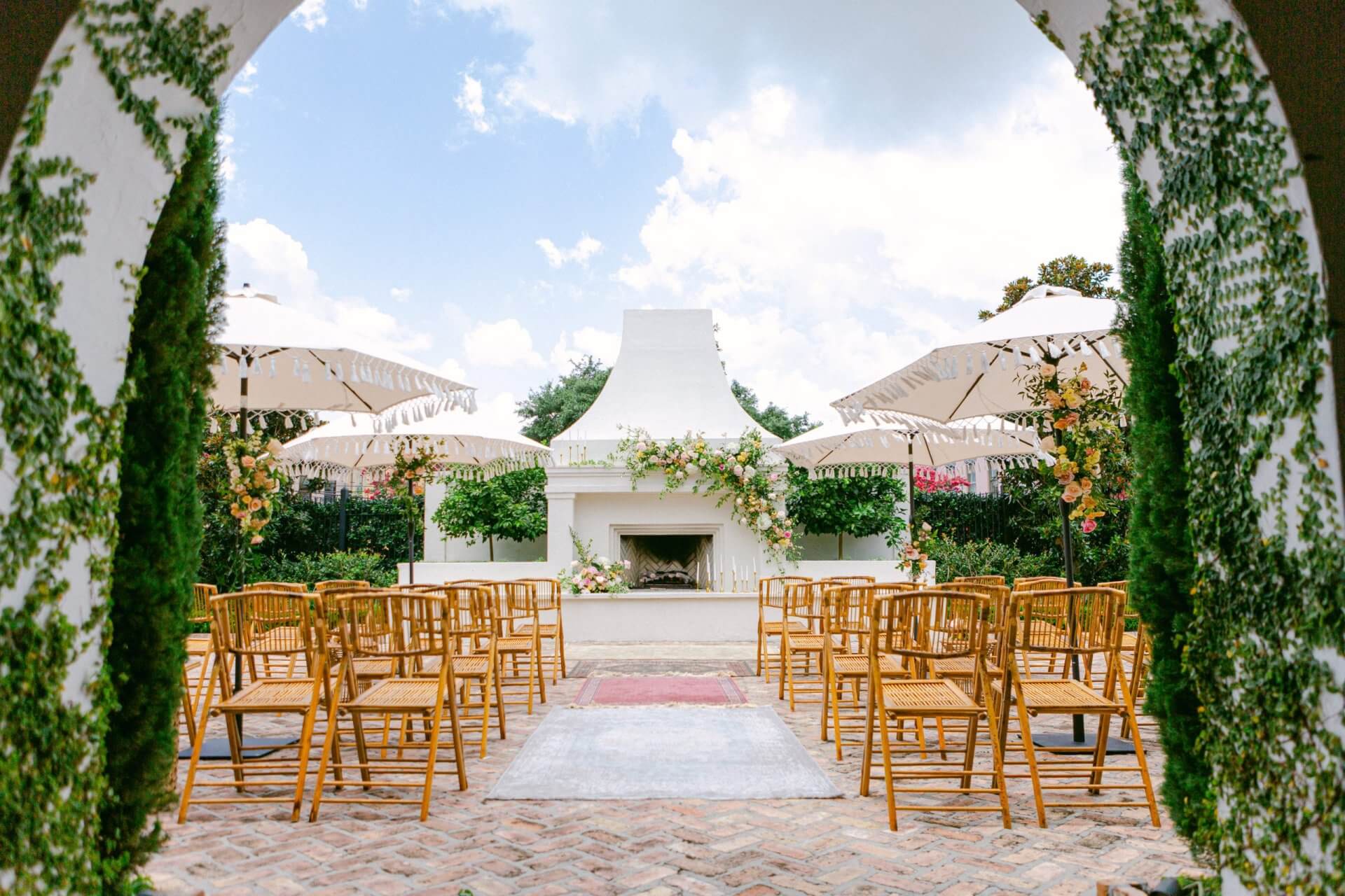 Outdoor wedding ceremony setup at one of the charming New Orleans wedding venues, featuring wooden chairs, umbrellas, and a flower-adorned white fireplace, viewed through an archway covered with lush greenery.