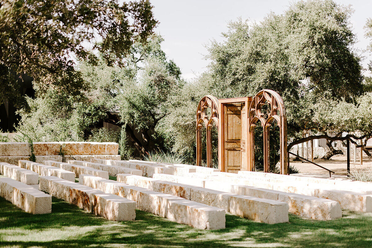 Outdoor stone seating area with rows of rectangular benches facing a decorative wooden arch and door structure, surrounded by trees and greenery—perfect for couples seeking unique San Antonio wedding venues.