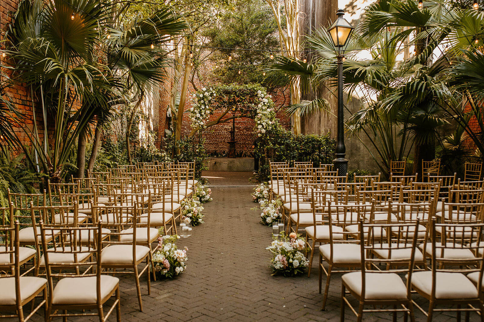 Rows of gold chairs surround an aisle lined with flower arrangements, leading to a floral wedding arch in an outdoor garden courtyard—perfect for couples seeking enchanting New Orleans wedding venues.