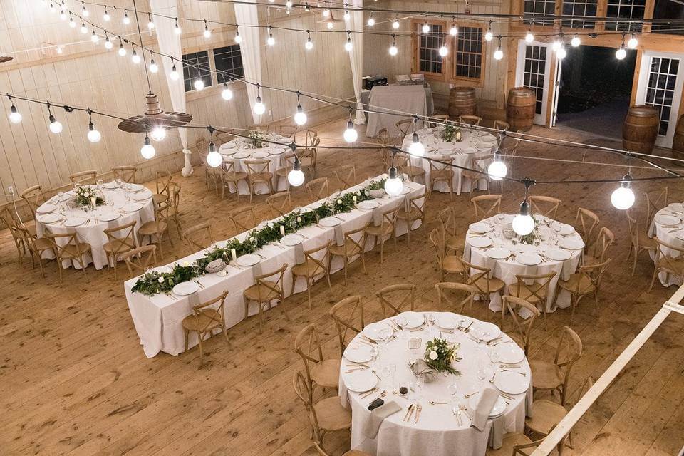 A decorated banquet hall with two long rectangular tables and four round tables set for dining, surrounded by wooden chairs and string lights overhead—one of the most charming wedding venues in Austin.