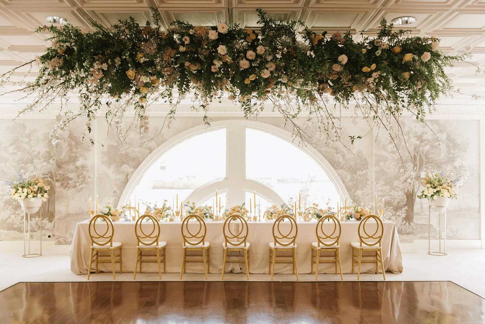 A long table with gold chairs is set for an event beneath a grand hanging floral arrangement, with natural light streaming through a large arched window—capturing the elegance found in top New Orleans wedding venues.
