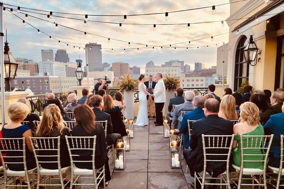 A wedding ceremony takes place on a rooftop at sunset, with guests seated in rows and the city skyline visible in the background—one of the most stunning scenes offered by New Orleans wedding venues.