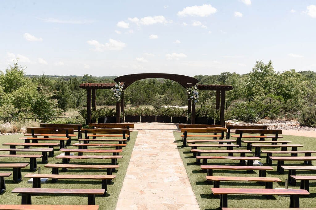Outdoor wedding ceremony setup with wooden benches, a stone aisle, and a floral-adorned wooden arch, surrounded by greenery and trees under a blue sky—perfect for couples seeking beautiful wedding venues in Fort Worth.